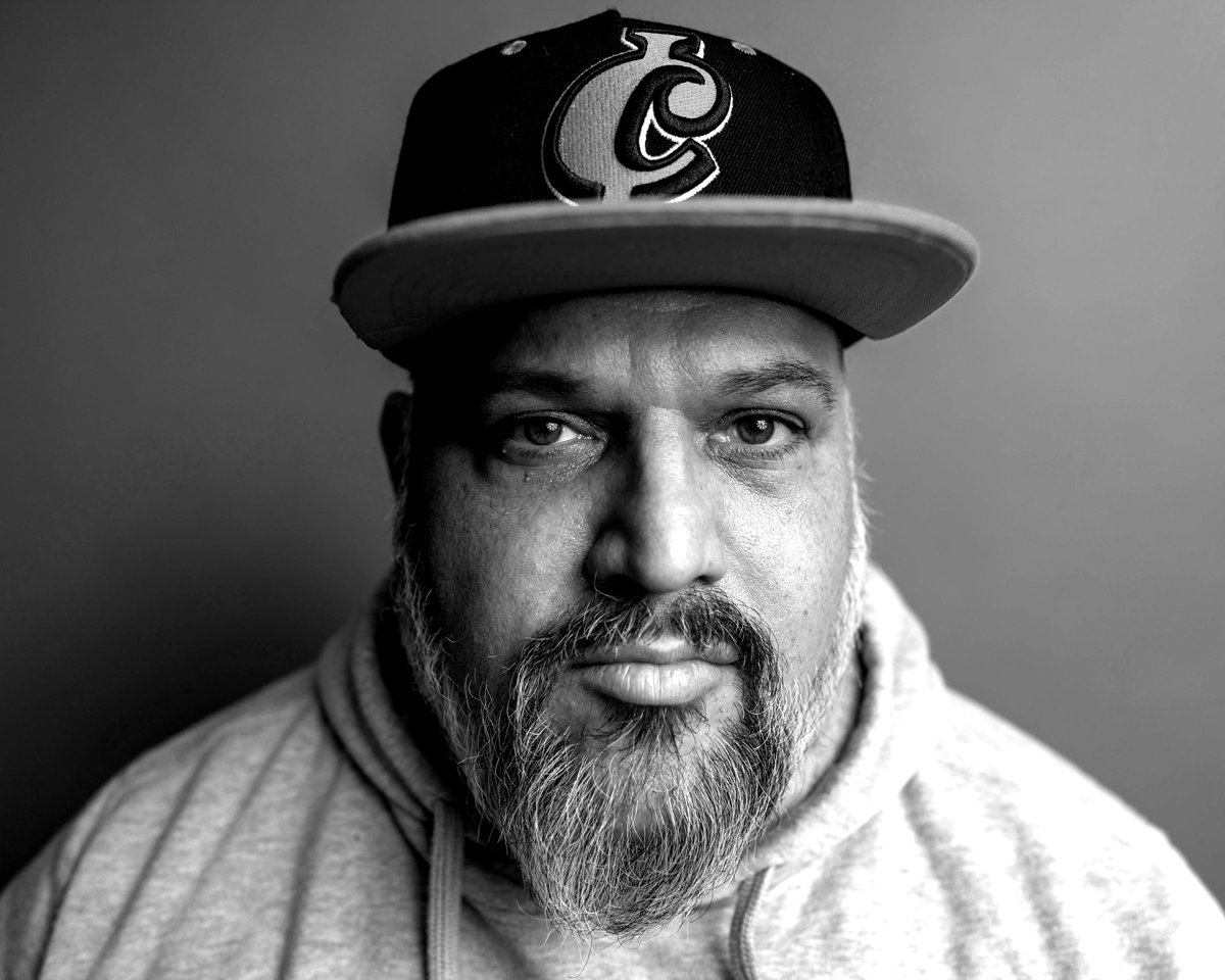 A high-contrast black-and-white portrait of Micah Baldwin: gray beard, baseball cap, gray hoodie, direct gaze.